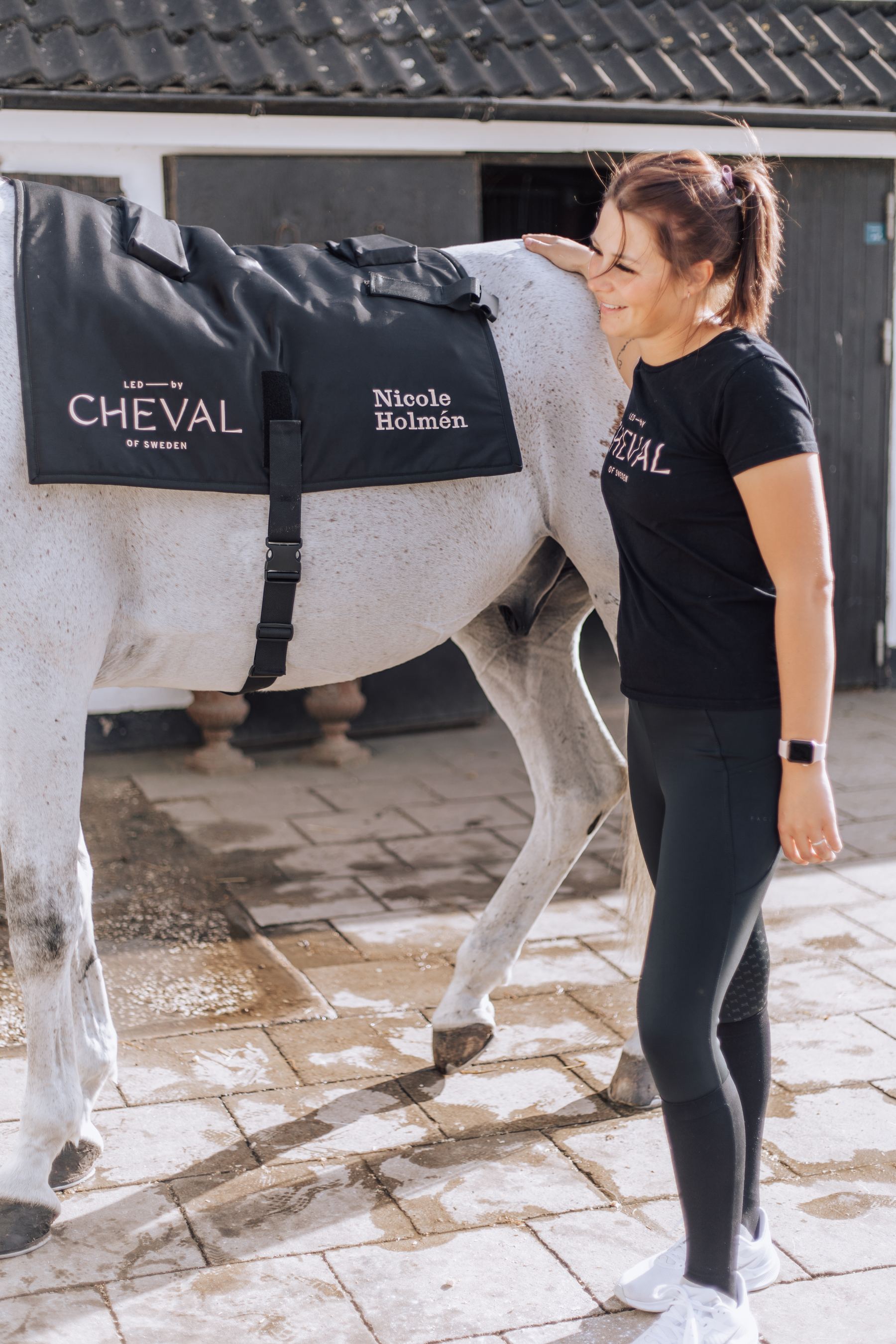 Nicole Holmén med sin grå häst på stallgården; hästen bär ett ” LED by CHEVAL ”-system